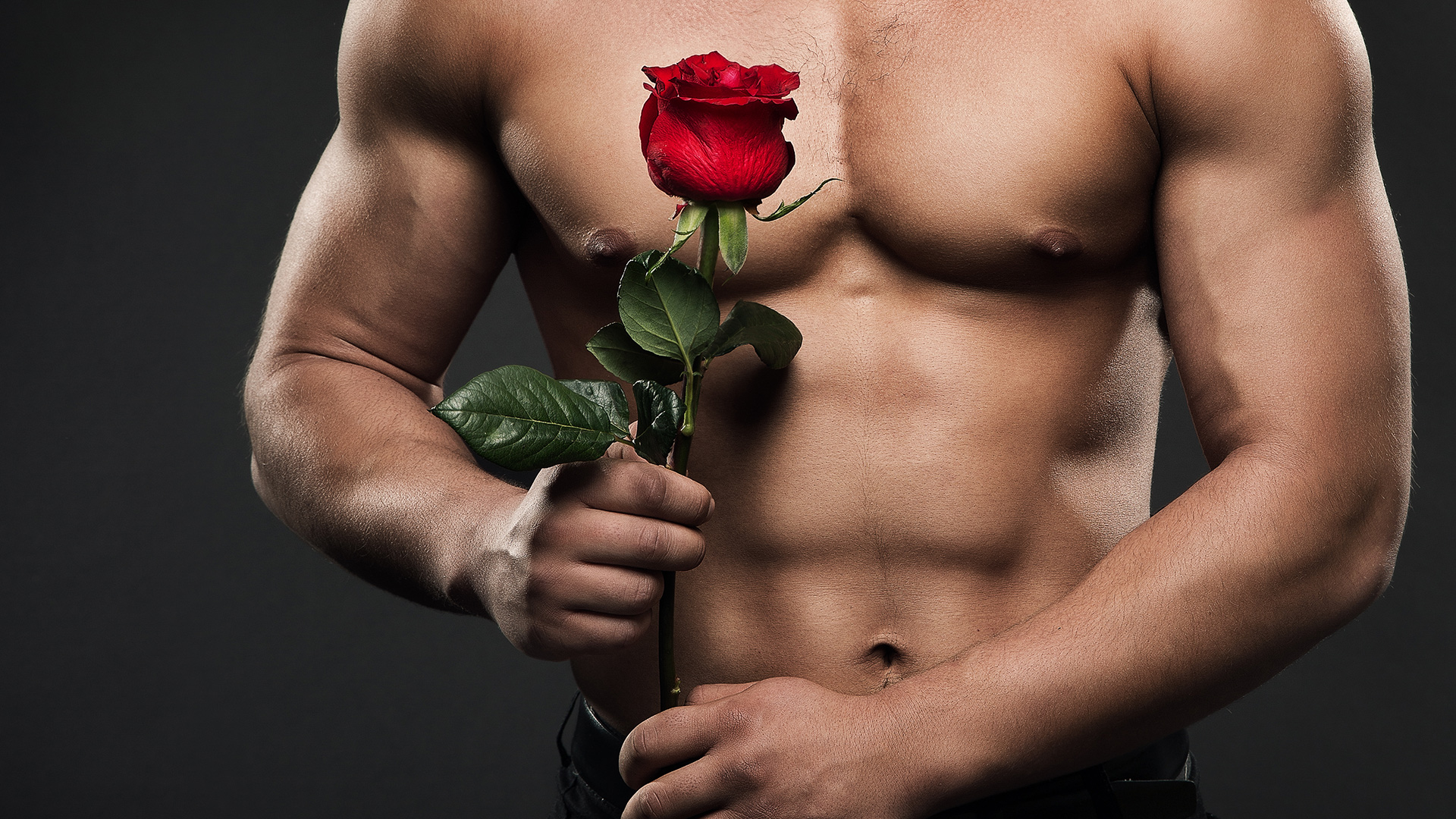 Shirtless upper body of a muscular man holding a red rose with both hands. Black background.